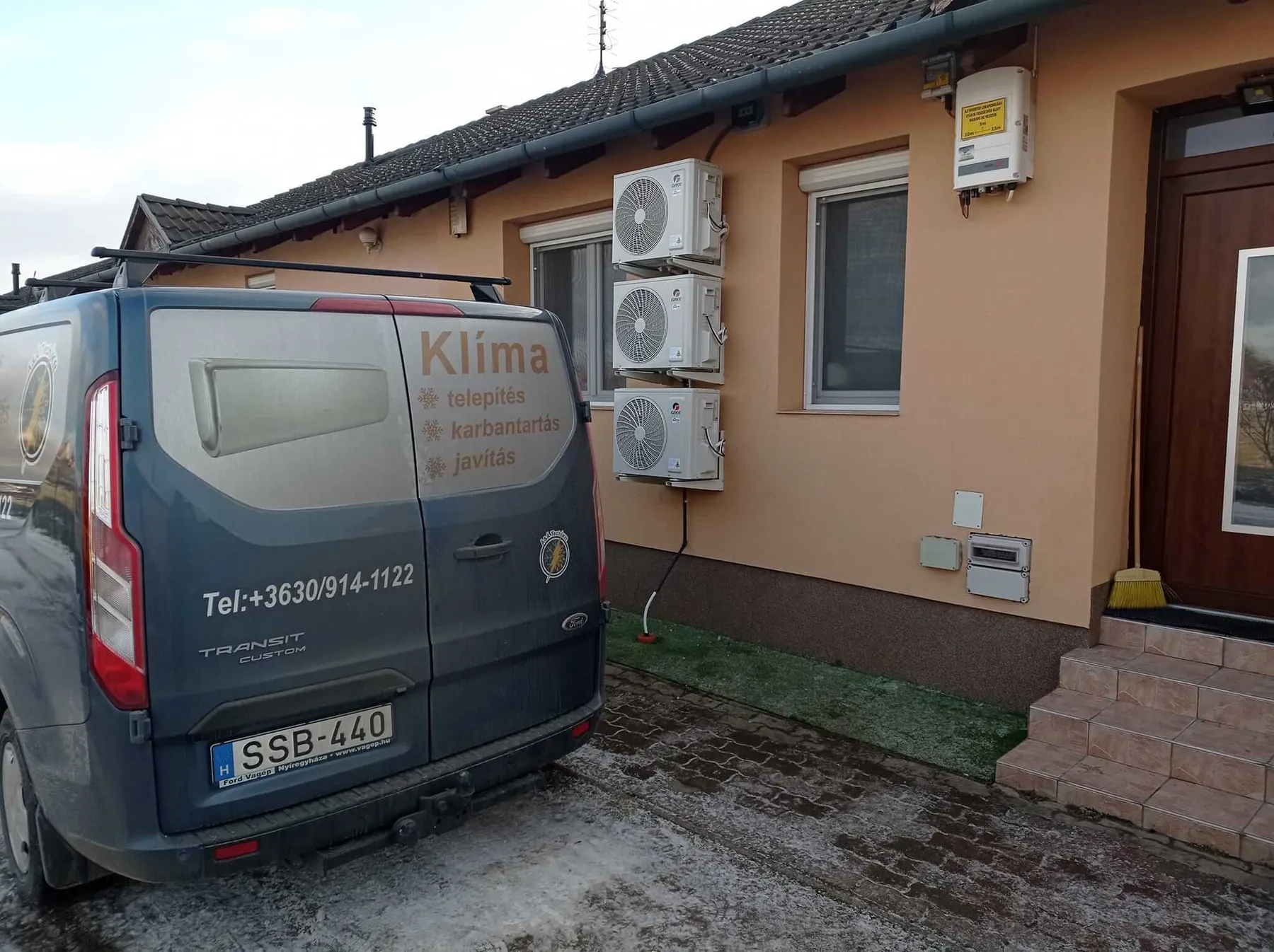 Kültéri klímaberendezések egy családi ház falán, a szakember furgonja és a bejárat látható.